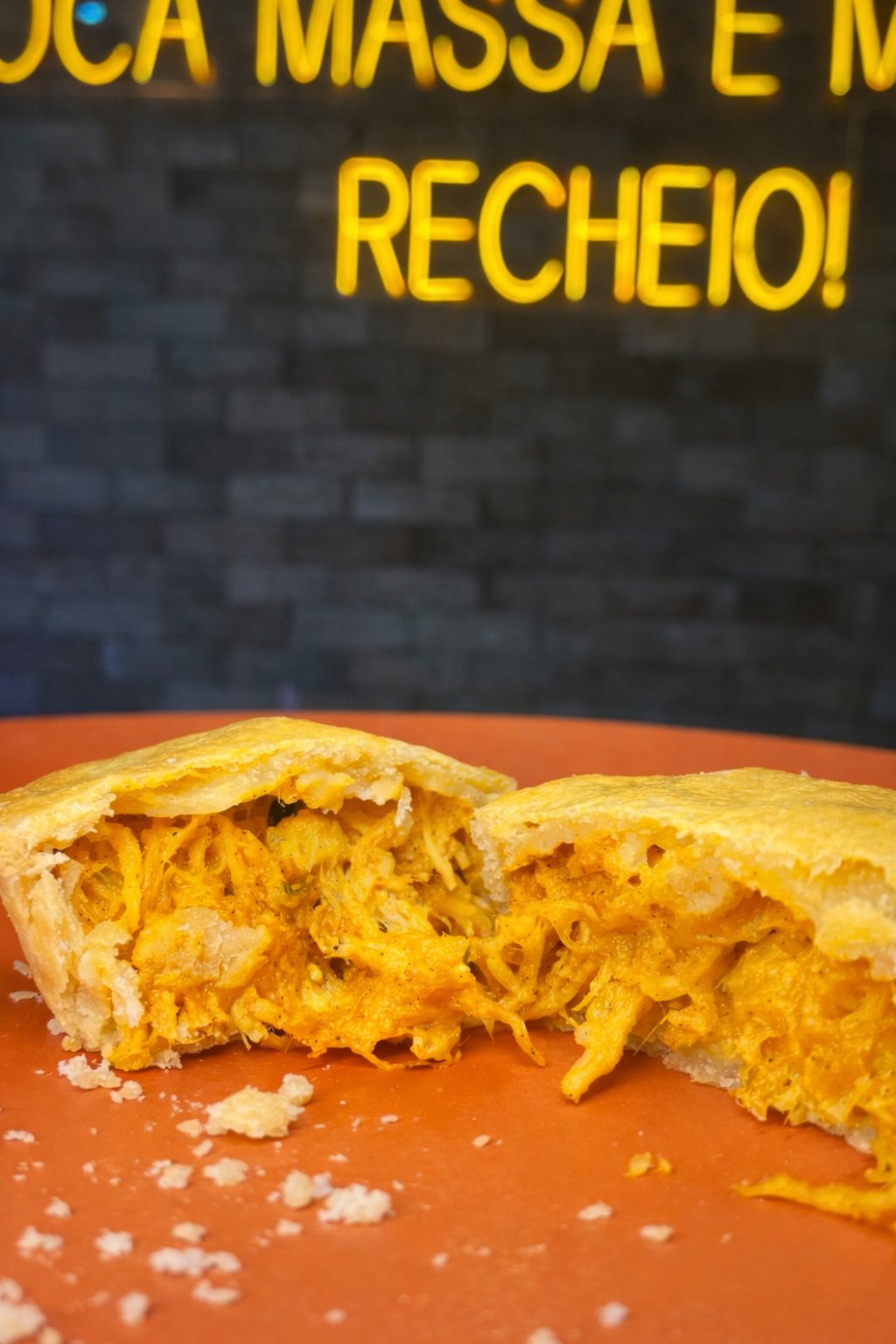 Empada de frango aberta ao meio com recheio fiapado generoso, diante do neon 'muito recheio' da Empada da Casa